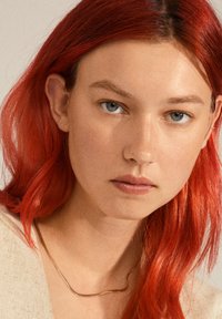Close-up of a person with red hair wearing a thin, wavy gold necklace against a neutral background, highlighting skin texture and eye color.