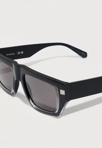 Mustat aurinkolasit, joissa on neliön muotoinen kehys, tummat liukuvärilliset linssit ja hillitty logodetalji sivussa. Mattapintainen, moderni muotoilu.