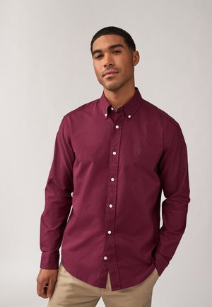 Camisa burdeos de botones y mangas largas, con botones blancos y un pequeño logo en el lado izquierdo del pecho, llevada sobre pantalones ligeros de color beige.