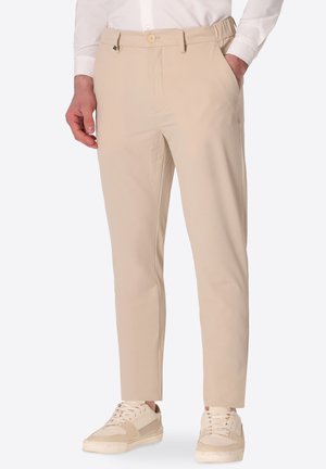 Uomo che indossa pantaloni beige aderenti, camicia bianca e sneakers beige, in piedi con una mano in tasca su sfondo bianco.