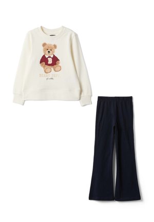 Cremefarbener Langarm-Sweatshirt mit Teddybär-Grafik und marineblauen ausgestellten Hosen auf weißem Hintergrund flach ausgelegt.
