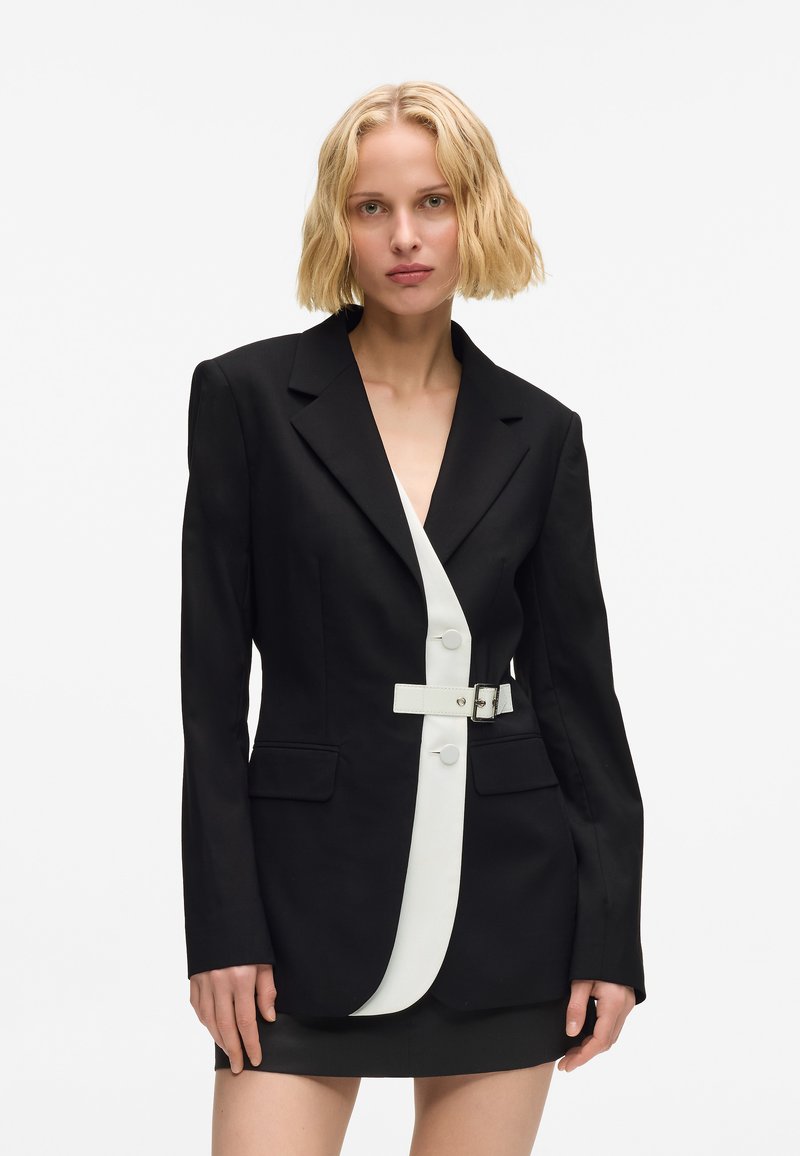 Melnā žakete ar strukturētu dizainu, balta centrālā panelis, pogas un jostu detaļa. Pielāgots siluets ar divām priekšējām kabatām, īss svārku garums.