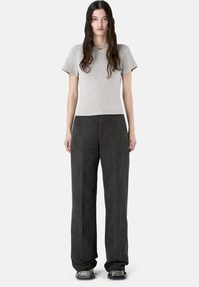 Kobieta stojąca z długimi ciemnymi włosami, ubrana w jasnoszary krótki top z wycięciem oraz luźne czarne spodnie i buty z kwadratowymi noskami.