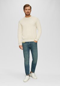 Pull crème avec un col rond, légère texture. Associé à un jean bleu slim et des baskets blanches, se tenant devant un fond uni.
