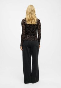 Juoda gėlių nėrinių ilgos rankovės palaidinė, dėvima ant prigludusio juodo drabužio, derinama su plačiais juodais džinsais, turinčiais matomus siuvinėjimo akcentus.