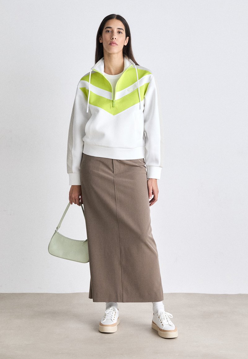 Weißer Sweatshirt mit grün-weißer Zickzack-Musterung, kombiniert mit einem langen braunen Rock. Hellgrüne Tasche und beige Plateau-Sneaker.