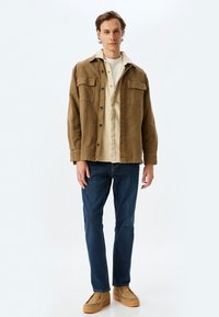 Chaqueta de camisa de pana marrón con dos bolsillos en el pecho sobre una camiseta beige, combinada con jeans de mezclilla azul y zapatos marrón claro.
