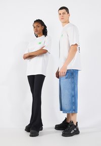 Two models wear casual clothing: a white t-shirt with a green graphic and black ribbed pants; the other wears a white tee and blue denim shorts.