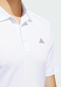 Bijela polo majica s ovratnikom, kratkim rukavima i dugmetima. Sadrži sivi Adidas logo na lijevoj strani prsnog područja. Glatka tekstura.