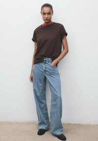Camiseta marrón de manga enrollada con un cuello rojo, combinada con jeans de tiro alto de un azul claro con una textura sutil. Zapatos negros que completan el conjunto.