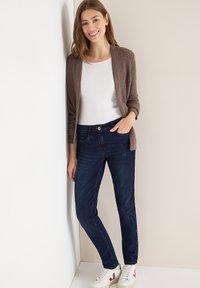 Ruskea ribbineuletakki valkoisen paidan päällä, yhdistetty tummansinisiin farkkuihin ja valkoisiin lenkkareihin. Malli seisoo neutraalia taustaa vasten.