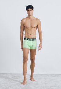 Boxeri de culoare verde neon cu un elastic negru pe care apare logo-ul "CALVIN KLEIN"; textură netedă; modelul stă pe o suprafață gri deschis.