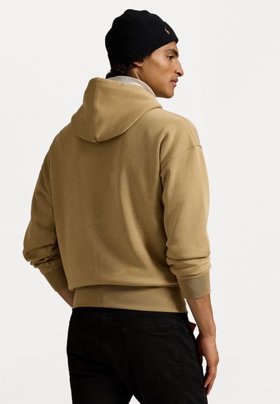 Sweatshirt bege com uma textura suave e capuz espaçoso, apresentando punhos e bainha canelados. O modelo usa um gorro preto de malha.