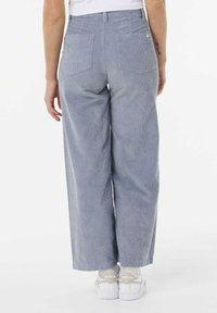 Pantaloni in corda a gamba larga di colore azzurro chiaro con strisce verticali, dotati di doppie tasche posteriori e una vestibilità alta in vita. Sneakers bianche visibili sotto.