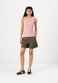 Hellrosa geripptes Tanktop, olivgrüne Shorts und schwarze Sneakers mit sandfarbenen Sohlen. Das Modell steht vor einem schlichten weißen Hintergrund.