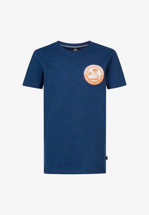 Dziecko noszące niebieską koszulkę z okrągłym pomarańczowym logo, jasnoniebieskie dżinsy i białe trampki, stojące z rękami w kieszeniach.