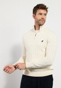 Cremefarbener Kabelstrickpullover mit hoher Kragen und Reißverschluss, gerippten Bündchen und Saum, mit einem kleinen Logo-Akzent auf der Brust.