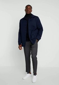 Man in een marineblauwe jas over een zwarte coltrui, grijs geruite broek en witte sneakers, staande tegen een effen lichte achtergrond.