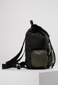 Sac à dos en tissu noir avec un dessus arrondi, des bretelles ajustables et une poche avant verte. Il dispose d'une fermeture à cordon et d'accents en zipper.