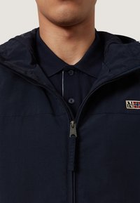 Man som bär en marinblå dragkedjejacka över en marinblå knäppt polo-skjorta, synlig lapplogo på jackans vänstra sida.