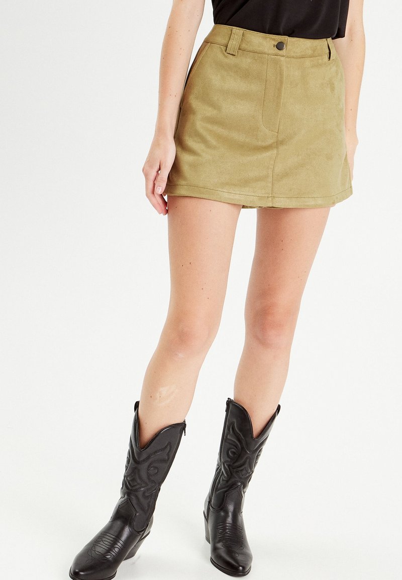 Olive green suede mini skirt with a high waist, front pockets, and a button closure, paired with black cowboy boots featuring embossing details.