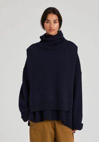 Kvinna som bär en oversize mörkblå polotröja och senapsfärgade byxor, stående mot en enkel ljus bakgrund.