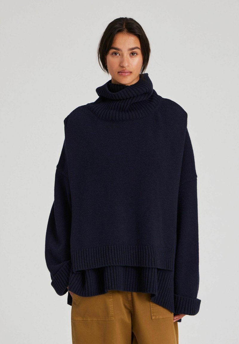 Kvinna som bär en oversize mörkblå polotröja och senapsfärgade byxor, stående mot en enkel ljus bakgrund.