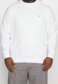 Sudadera blanca hecha de un tejido suave con un bolsillo delantero, cordón ajustable y un pequeño logo en el pecho. Combinada con pantalones a rayas.
