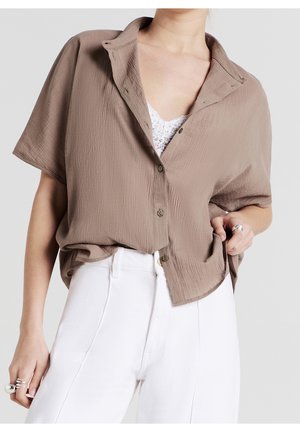 Femme portant une chemise marron ample à manches courtes boutonnée par-dessus un top en dentelle blanc avec un pantalon blanc taille haute et des bagues argentées aux deux mains.