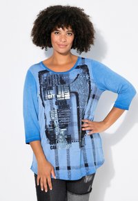 T-shirt à manches longues bleu avec du texte graphique et des motifs dans différentes nuances de bleu, présentant un col rond et une coupe décontractée.