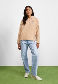 Khaki fleece pullover hoodie with front pocket and drawstring hood, paired with light blue straight-leg jeans and cream sneakers.