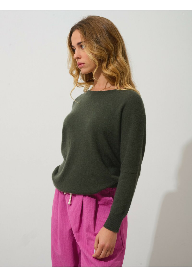 Olivgrüner Strickpullover mit rundem Halsausschnitt, lockerer Passform und langen Ärmeln, kombiniert mit pinken, plissierten Hose mit Kordelzug.