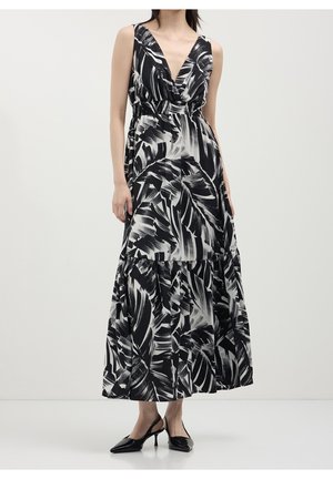 Kobieta ubrana w czarno-białą maxi sukienkę bez rękawów z wzorem liści oraz czarne szpiczaste buty na obcasie typu slingback.