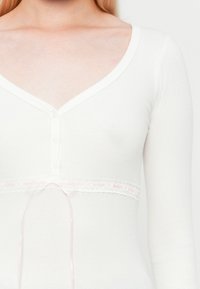Weißes, geripptes Langarmoberteil mit tiefem V-Ausschnitt, das einen geknöpften Vorderteil und ein dekoratives Spitzenband mit einem rosa Band in der Taille aufweist.