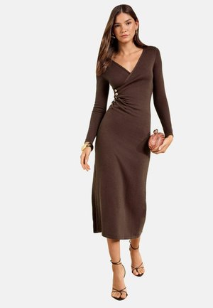 Femme portant une longue robe marron ajustée avec des boutons sur le côté, tenant une pochette ronde en bois, et portant des talons noirs à brides et des bijoux en or.