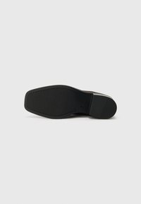 Musta kumipohja, jossa on teksturoitu pinta ja vahvistettu kanta, which has a smooth edge and minimal branding, valkoisella taustalla.