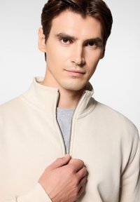 Jasno beżowa bluza na zamek z wysokim kołnierzem, o miękkiej fakturze, z detalem srebrnego zamka, noszona na szarym t-shircie.