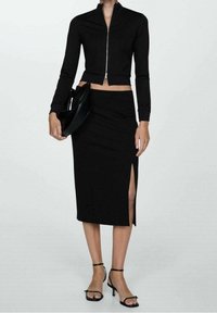 Sort to-delt tøj sæt bestående af en cropped, langærmet zip-jakke og en tætsiddende nederdel med en sideslids. Accentueret med strappy sorte hæle og en taske.