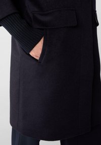 Marineblauer Mantel aus strukturiertem, weichem Stoff mit gerippten Bündchen und einer Seitentasche, der eine gerade Silhouette und minimalistische Details zeigt.