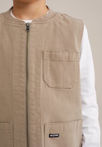 Beige ärmlös väst i canvas med dragkedja fram, som har två framfickor och en liten svart etikett vid nederkanten.