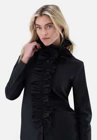 Black button-up shirt featuring a prominent ruffled front detail, smooth texture, and long sleeves with buttoned cuffs.