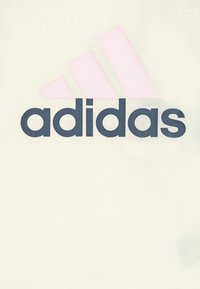 T-shirt di cotone chiaro con un grande logo "adidas" blu navy e tre strisce rosa sopra, creando un design grafico semplice.