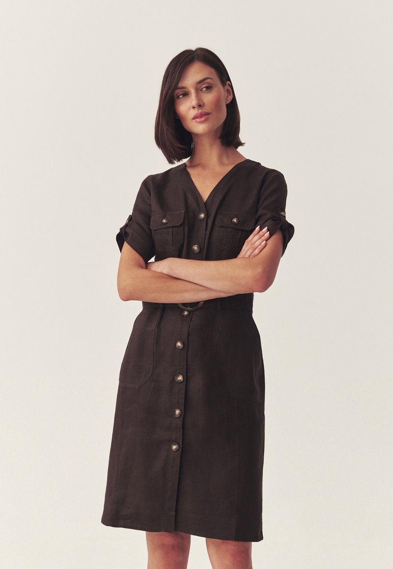 Femme aux cheveux courts et foncés portant une robe marron boutonnée avec manches retroussées, debout les bras croisés devant un fond uni.