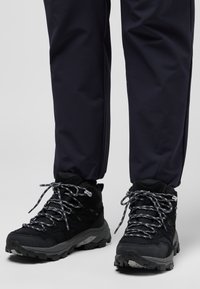Black hiking boots with textured synthetic material, silver speckled laces, and a rugged sole, paired with navy pants.