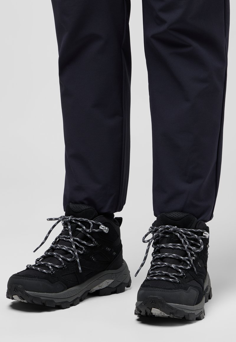 Black hiking boots with textured synthetic material, silver speckled laces, and a rugged sole, paired with navy pants.