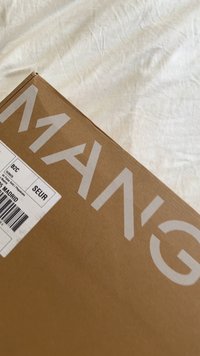 Hnědá kartonová přepravní krabice s částí textu „AMAZING“ a bílou přepravní štítkem na bílé látkové podložce.