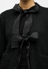 Černý pletený cardigan s texturovaným povrchem, otevřeným předním dílem a saténovou mašlí u krku. Nositý přes černý žebrovaný top.