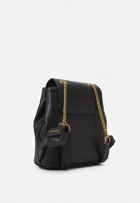 Černý kožený batoh se strukturovaným tvarem, zlatými řetízkovými akcenty a malým koženým štítkem s názvem značky. Hladký povrch.