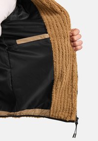 Chaqueta de felpa color beige con textura acanalada en el exterior. El forro de satén negro cuenta con un bolsillo beige. Mano sosteniendo la chaqueta.