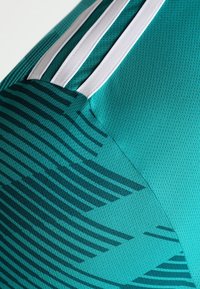 Teal sportovní tkanina s tmavými geometrickými pruhy a třemi bílými paralelními pruhy na texturovaném materiálu, ukazující detail ramene oděvu.